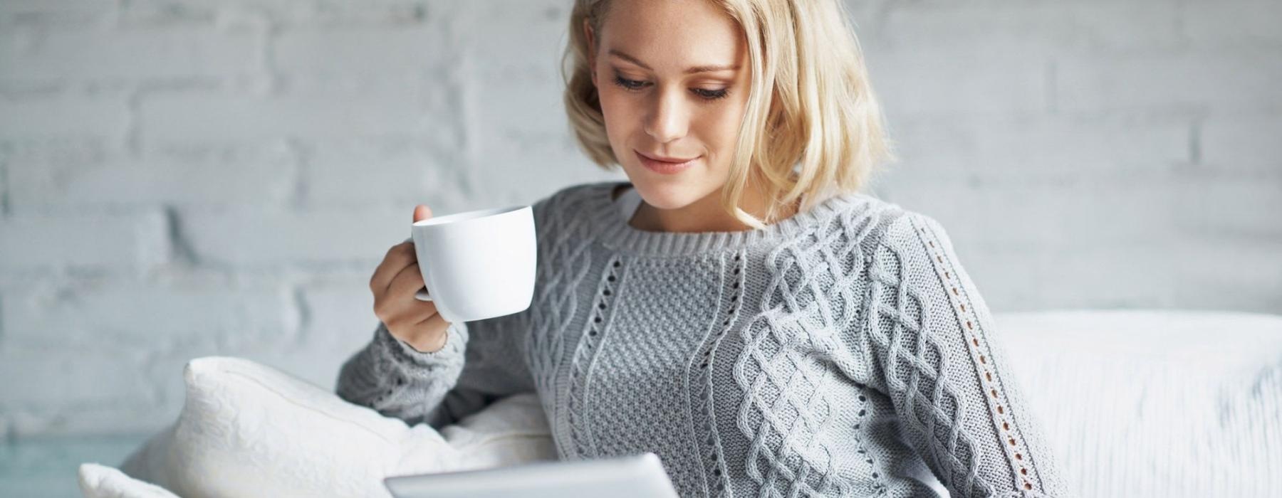 a woman sits on a couch with a cup of coffee and looks at her tablet